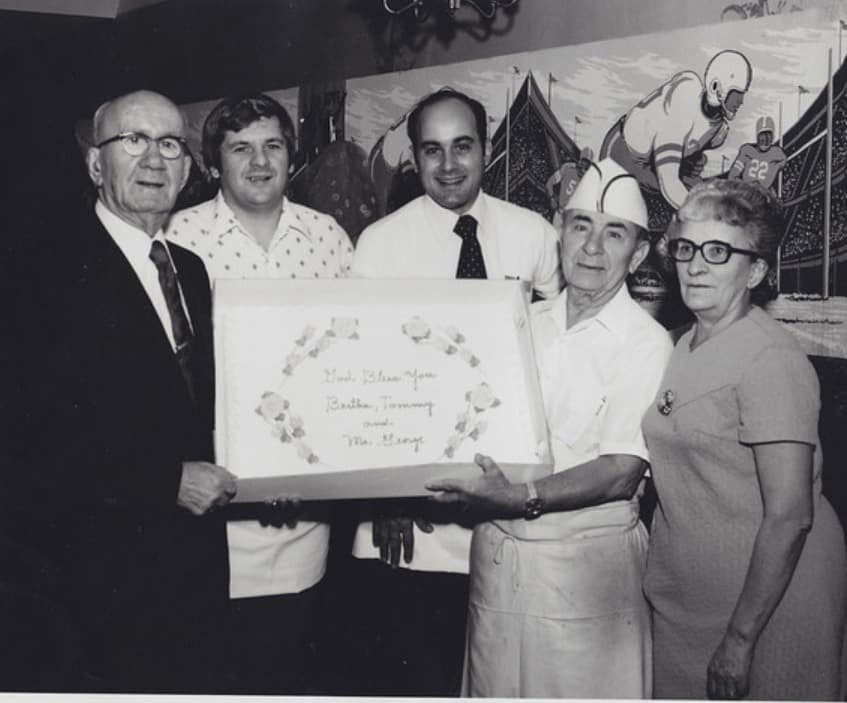 Black-and-white photo of five people holding a cake decorated with a floral wreath and the handwritten message “God Bless You, Bertha, Tommy and Mr. George,” in front of a football-themed mural