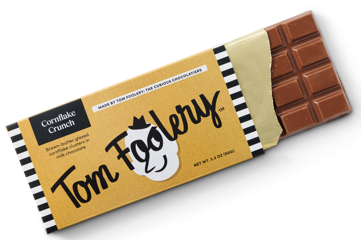 A single wrapped chocolate bar on a clean white background. The wrapper is tan with black-and-white striped edges, large 'Tom Foolery' script, and 'Cornflake Crunch' text.
